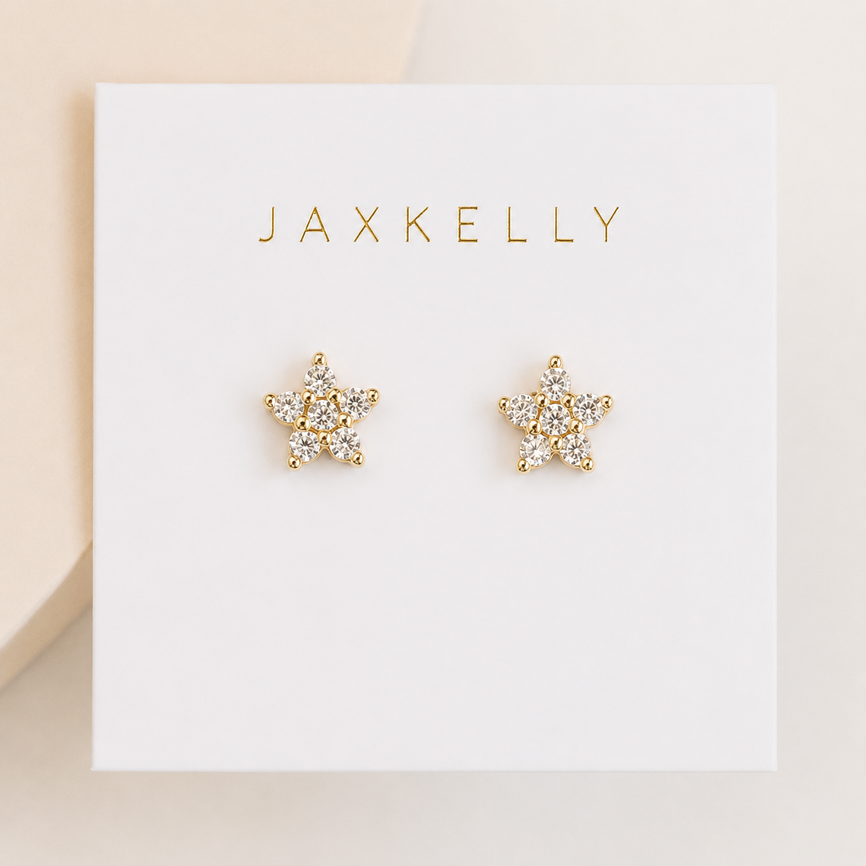 JaxKelly Pavé Flower earrings feature gold flower-shaped studs with sparkling crystals and sterling silver posts, presented on a white card with JaxKelly in gold lettering at the top.