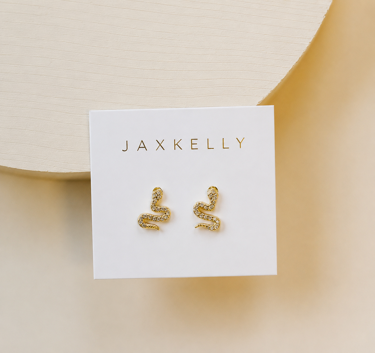 The JaxKelly Pavé Snake earrings—gold plated and adorned with sparkling CZ stones—are displayed on a white JaxKelly card against a light beige background.