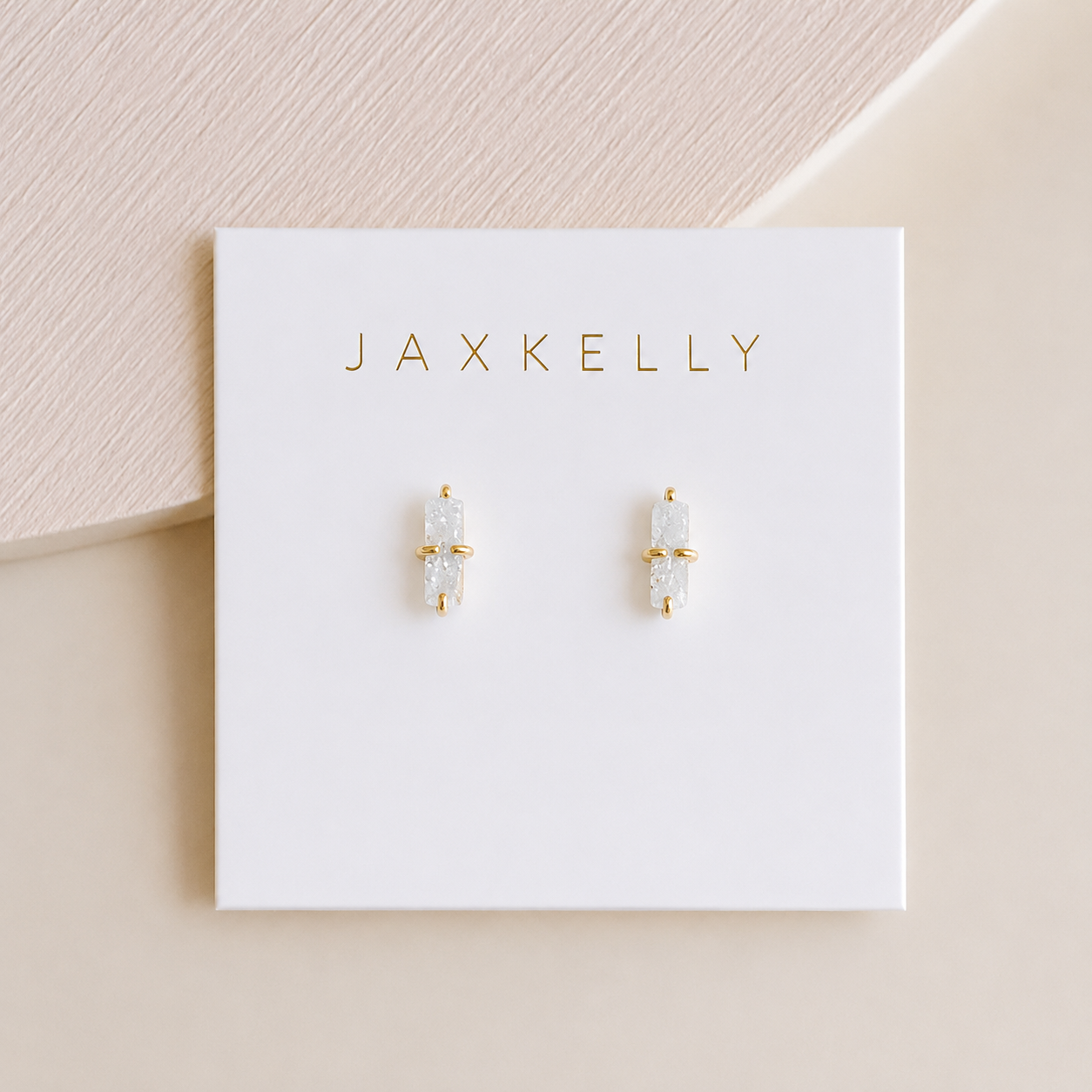 A pair of small, hypoallergenic 18K gold plated White Bar Druzy earrings by JaxKelly with clear natural druzy stone studs, displayed on a white JaxKelly card. Soft beige and cream tones in the background.