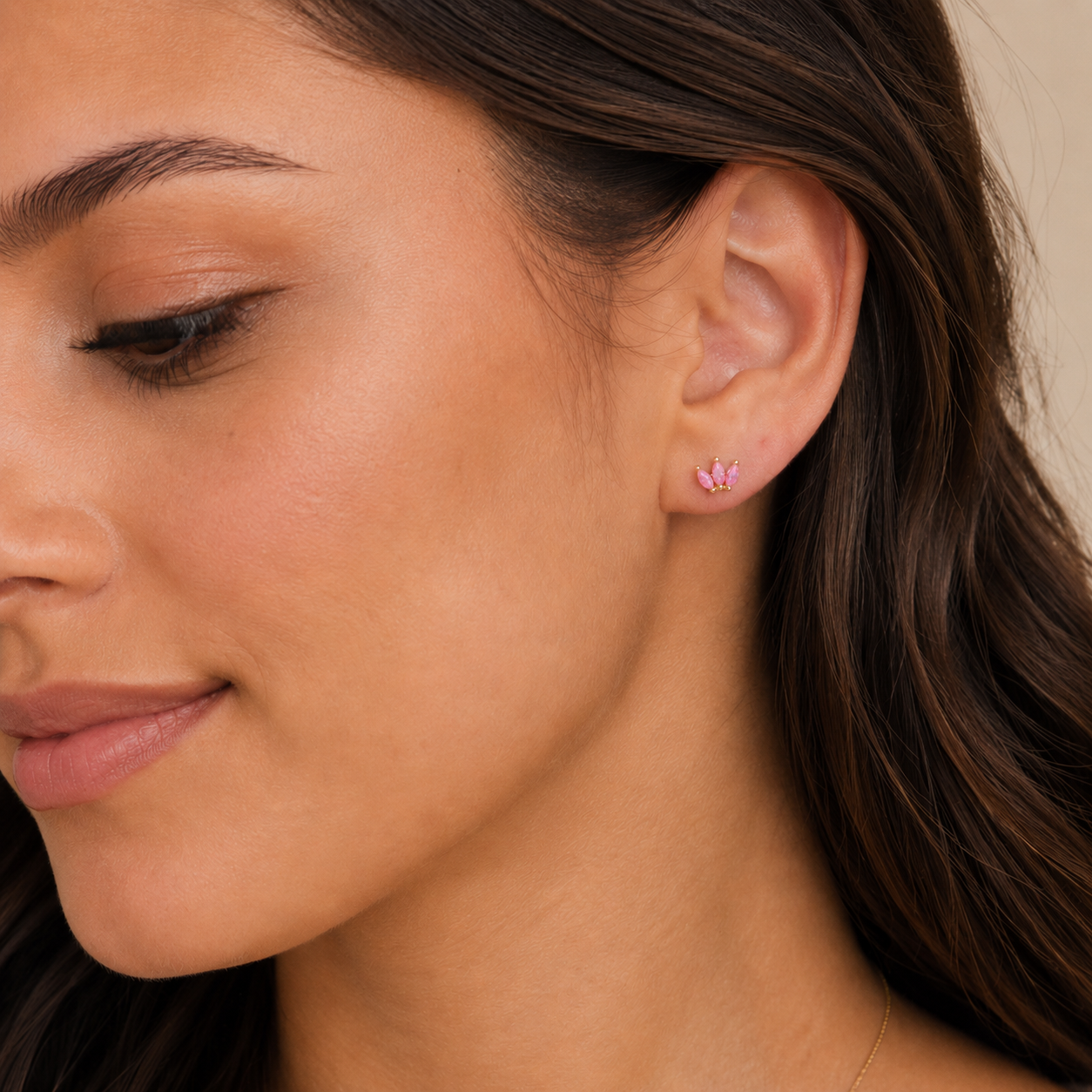 A close-up of a woman with long brown hair wearing JaxKelly’s Pink Opal Crown—dainty flower-shaped lab-grown gemstone stud earrings set in 18k gold plating. She has smooth skin and a gentle, natural expression.