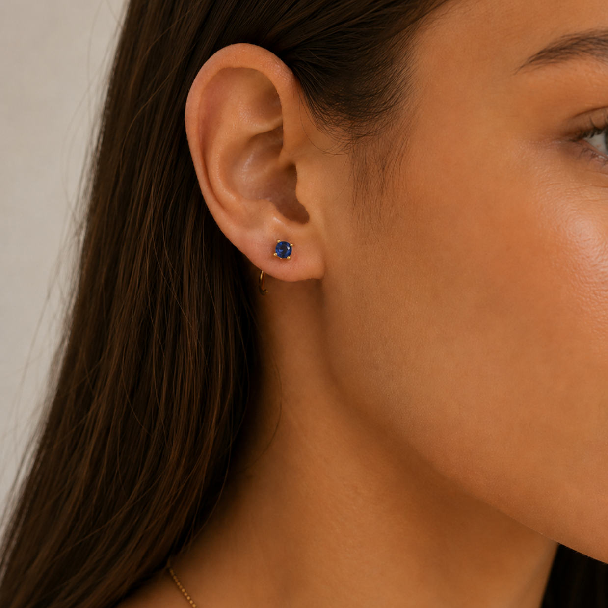 Close-up of a woman’s ear wearing JaxKelly’s Sapphire CZ Huggie and a small blue gemstone stud. Her straight brown hair is tucked behind her ear, and her skin appears smooth and radiant.