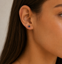 Close-up of a woman’s ear wearing JaxKelly’s Sapphire CZ Huggie and a small blue gemstone stud. Her straight brown hair is tucked behind her ear, and her skin appears smooth and radiant.