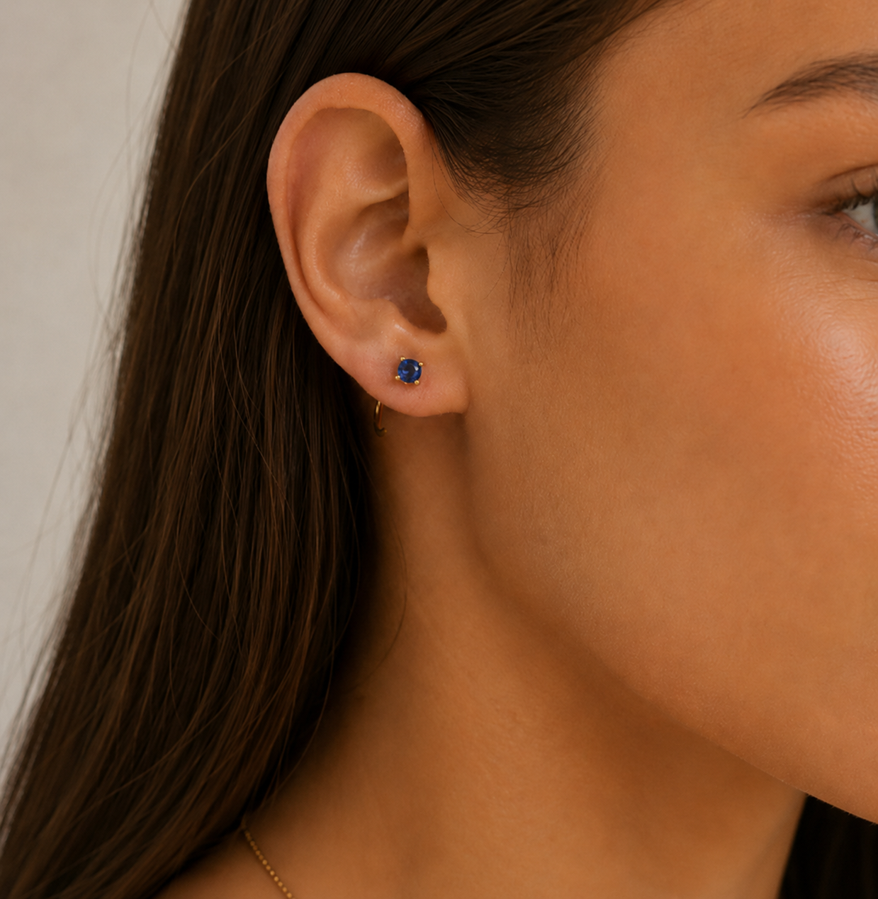 Close-up of a woman’s ear wearing JaxKelly’s Sapphire CZ Huggie and a small blue gemstone stud. Her straight brown hair is tucked behind her ear, and her skin appears smooth and radiant.