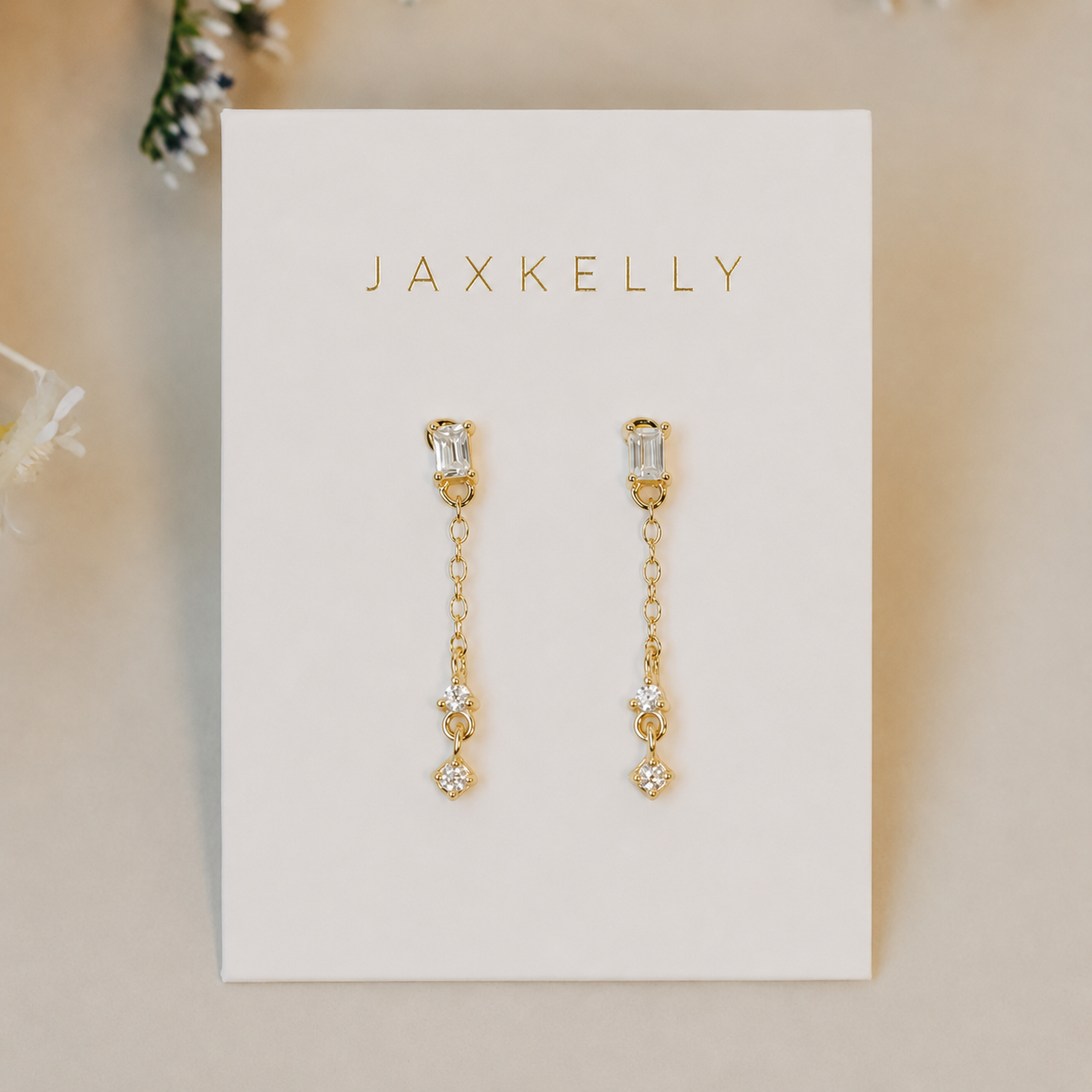 The JaxKelly Baguette Drop earrings feature 18k gold plating with rectangular and round gemstone accents, shown on a white JaxKelly card with small white flowers in the background.