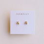 The JaxKelly Double Stud Stack features a pair of small, geometric 18k gold-plated stud earrings with a minimalist design and durable, protective coating. Displayed on a white card with JAXKELLY printed in gold at the top.