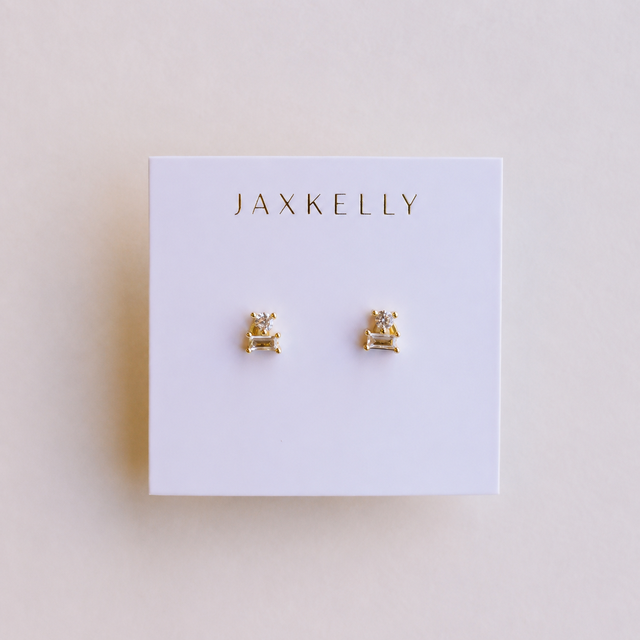 The JaxKelly Double Stud Stack features a pair of small, geometric 18k gold-plated stud earrings with a minimalist design and durable, protective coating. Displayed on a white card with JAXKELLY printed in gold at the top.