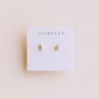 A pair of small Offset Trio - White CZ 18k gold plated stud earrings from JaxKelly is displayed on a white card with the brand name printed in gold at the top, set against a light beige background.