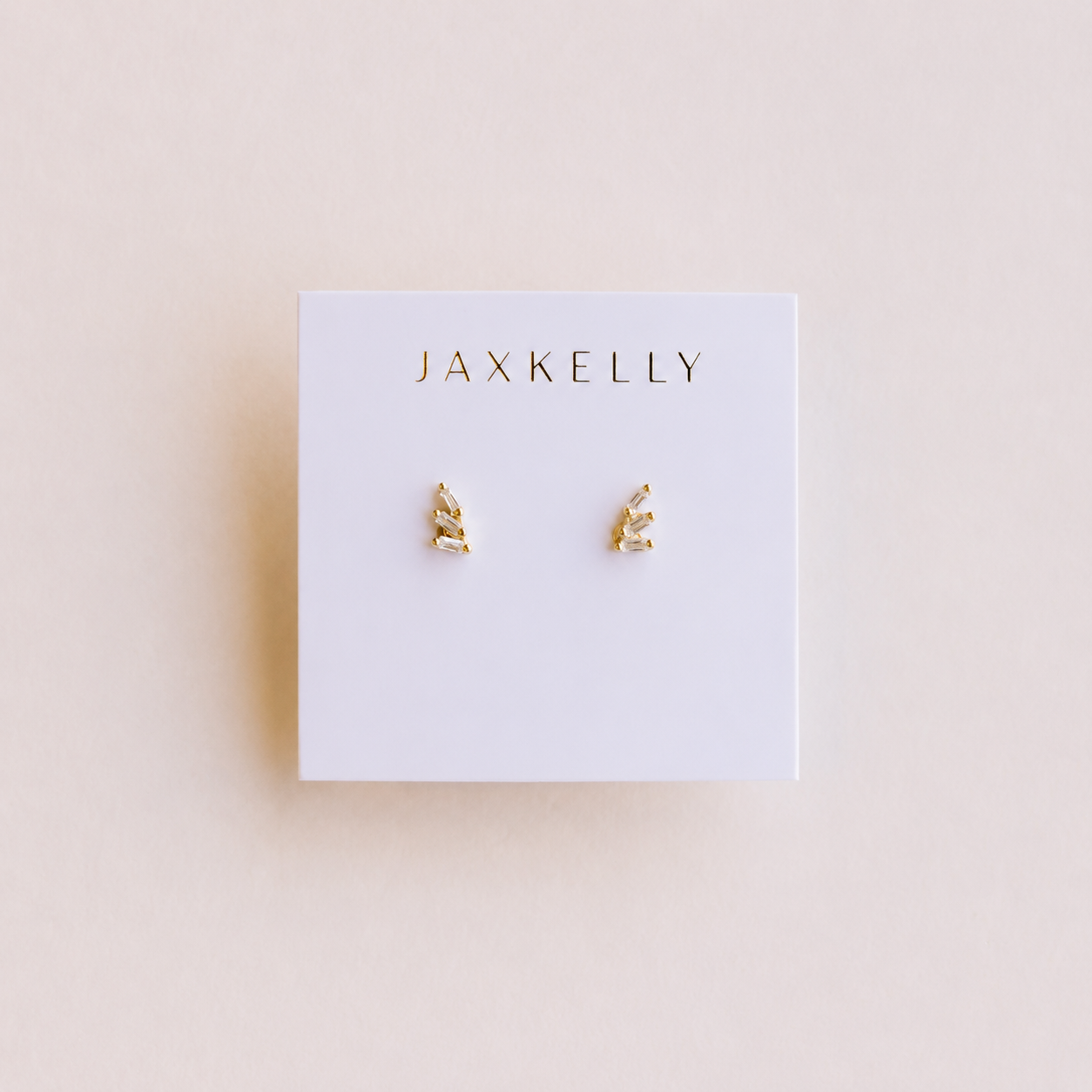 A pair of small Offset Trio - White CZ 18k gold plated stud earrings from JaxKelly is displayed on a white card with the brand name printed in gold at the top, set against a light beige background.