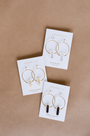 Three pairs of crystal earrings—Clear Quartz Hoop - Healing by JaxKelly, plus citrine and smoky quartz designs—are shown on white JaxKelly cards against a brown background. Each pair features beautiful stones on elegant sterling silver hoops.