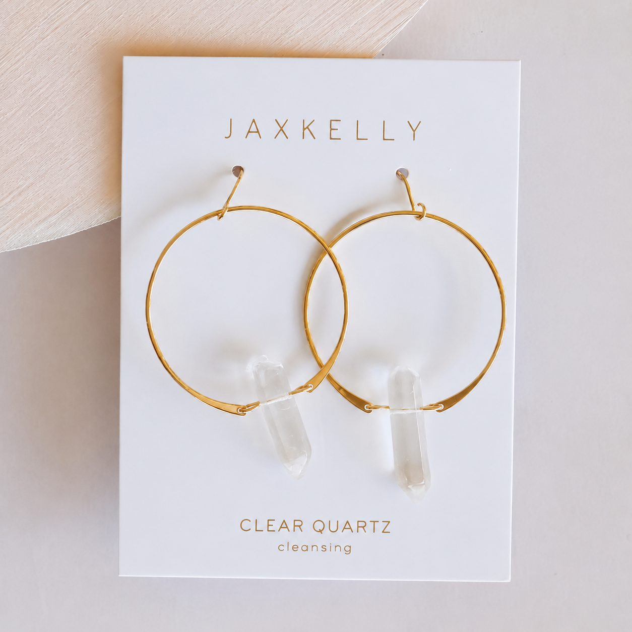 A pair of JaxKelly Clear Quartz Hoop - Healing earrings is displayed on a white card labeled JaxKelly, Clear Quartz, cleansing, resting on a light-colored surface.