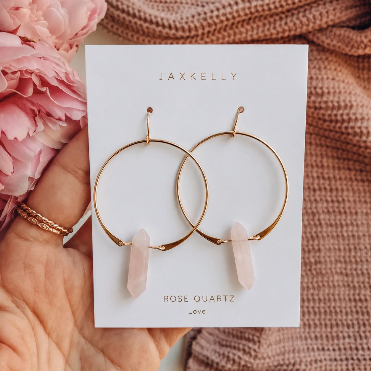A hand holds a card showing JaxKelly's Rose Quartz Hoop - Love earrings. Pink flowers and a knitted fabric are in the background.