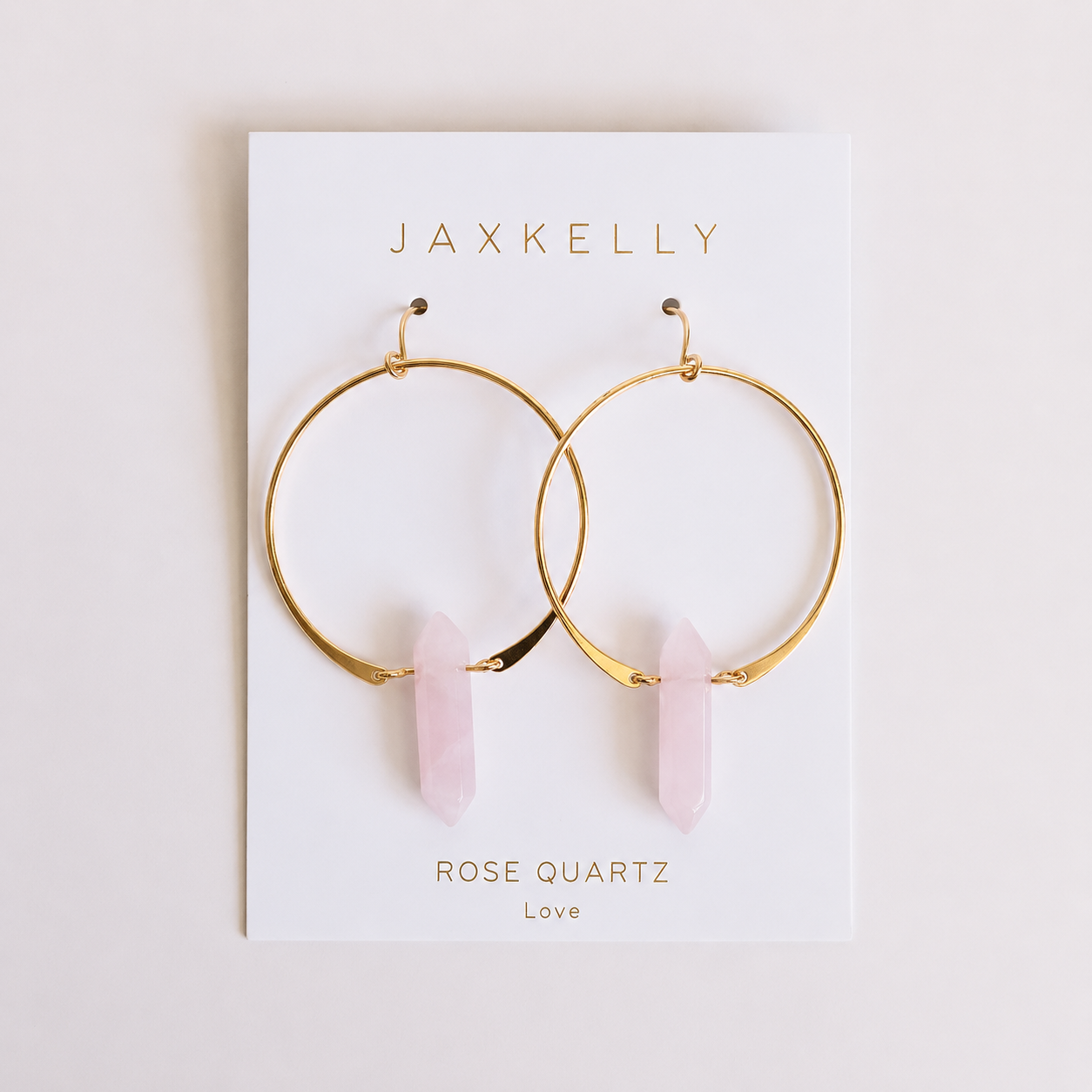 The JaxKelly Rose Quartz Hoop - Love earrings feature gold plated hoops with rose quartz crystal pendants, displayed on a white card with gold “JAXKELLY” and “ROSE QUARTZ Love” text.