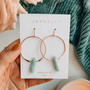 A hand holds a JaxKelly card displaying the Amazonite Hoop - Hope earrings. The card reads "AMAZONITE hope." A pink rose and teal textured fabric appear in the background.