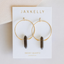 A pair of large gold crystal hoops with dark smoky quartz pendants from JaxKelly, displayed on a white card labeled "Smoky Quartz Hoop - Protection"—a stylish piece of handcrafted jewelry.