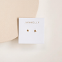 A white card with “JaxKelly” in gold displays a pair of Tiny Star mini stud earrings with 18k gold-plated sterling silver posts, set on a soft beige background and accented by a wooden circle in the upper left corner.
