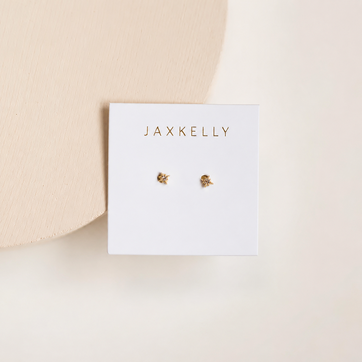 A white card with “JaxKelly” in gold displays a pair of Tiny Star mini stud earrings with 18k gold-plated sterling silver posts, set on a soft beige background and accented by a wooden circle in the upper left corner.