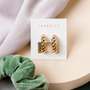The Twisted Hoop by JaxKelly features 18k gold plating and sterling silver posts, presented on a white JaxKelly card atop a beige surface with a green scrunchie and lilac fabric nearby.