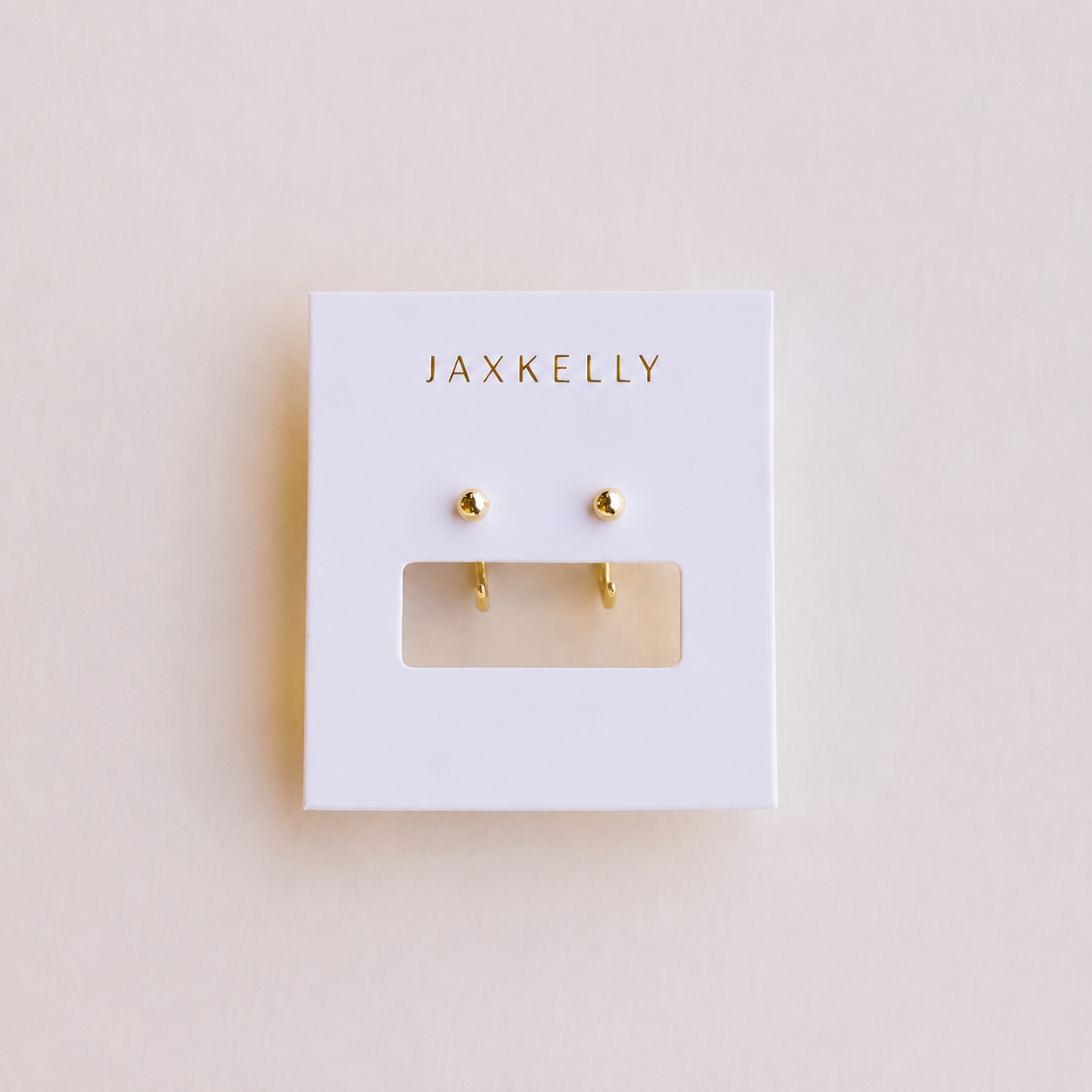 A pair of Gold Sphere Huggie earrings by JaxKelly displayed on a white, tarnish-free card with "JaxKelly" printed in gold at the top and a rectangular cutout beneath the earrings.