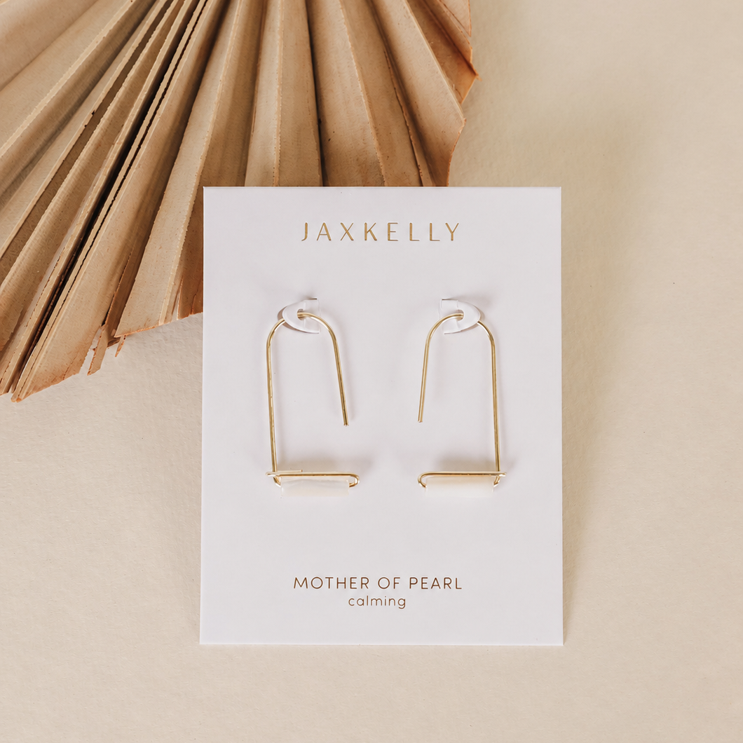 A pair of JaxKelly Mother of Pearl Gemstone Drop - Calming earrings is displayed on a branded card, set against a beige background with a dried palm leaf in the upper left corner.
