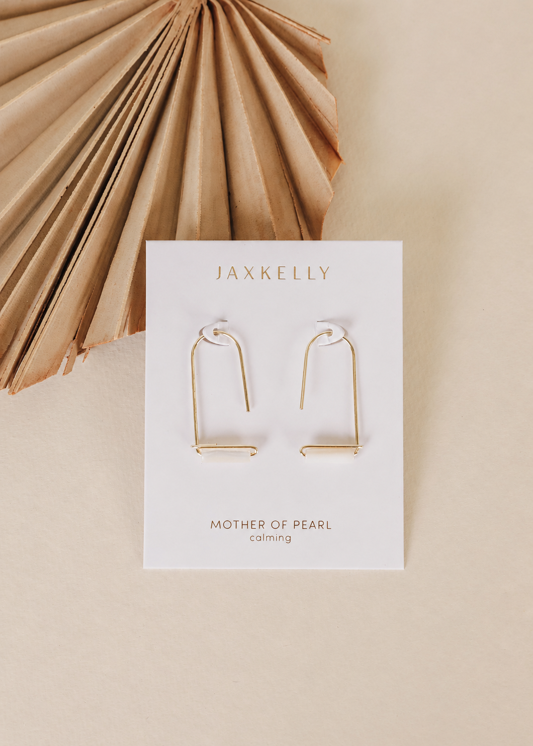 A pair of JaxKelly Mother of Pearl Gemstone Drop - Calming earrings is displayed on a branded card, set against a beige background with a dried palm leaf in the upper left corner.