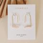 A pair of gold-plated JaxKelly Rose Quartz Gemstone Drop - Love earrings with horizontal rose quartz stones, shown on a branded ROSE QUARTZ "love" card, set against a neutral background with a dried palm leaf.