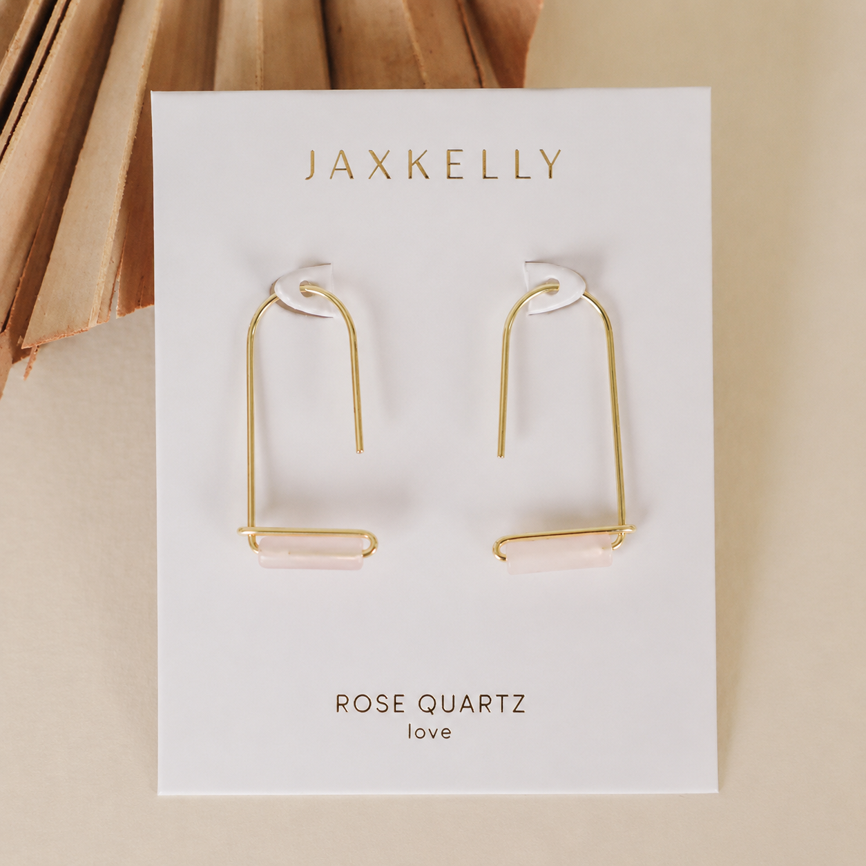 A pair of gold-plated JaxKelly Rose Quartz Gemstone Drop - Love earrings with horizontal rose quartz stones, shown on a branded ROSE QUARTZ "love" card, set against a neutral background with a dried palm leaf.