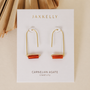 The JaxKelly Carnelian Agate Gemstone Drop - Creativity earrings feature 18k gold plating and horizontal carnelian agate stones, displayed on a white branded card against a neutral background with dried palm leaves.