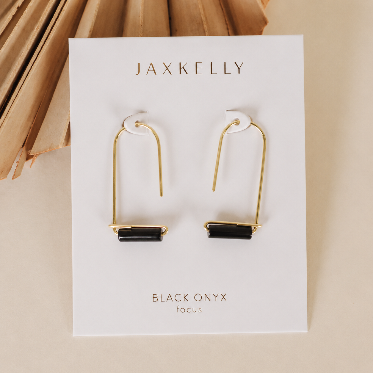 A pair of JaxKelly Black Onyx Gemstone Drop - Focus earrings with gold plating and rectangular black onyx stones, displayed on a branded white card, rests on a beige surface with a decorative accent in the corner.