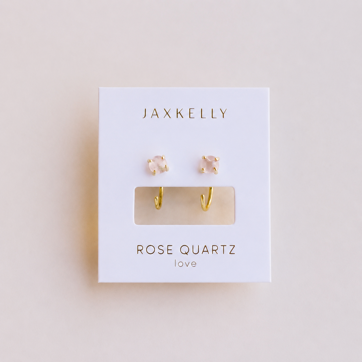 A pair of elegant Rose Quartz Huggie earrings by JaxKelly, showcased on a white card labeled "ROSE QUARTZ love," with the stylish gold huggie hoops and pink stones visible through a clear window.