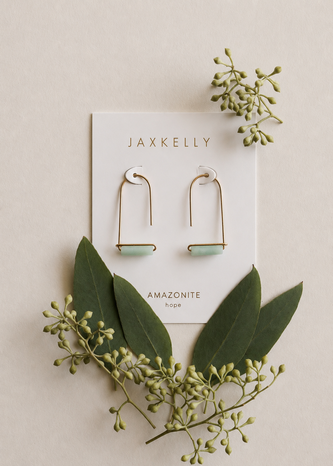 A pair of Amazonite Gemstone Drop - Hope earrings by JaxKelly, 18k gold plated, displayed on a white card reading "AMAZONITE hope," surrounded by green leaves and sprigs on a beige background.