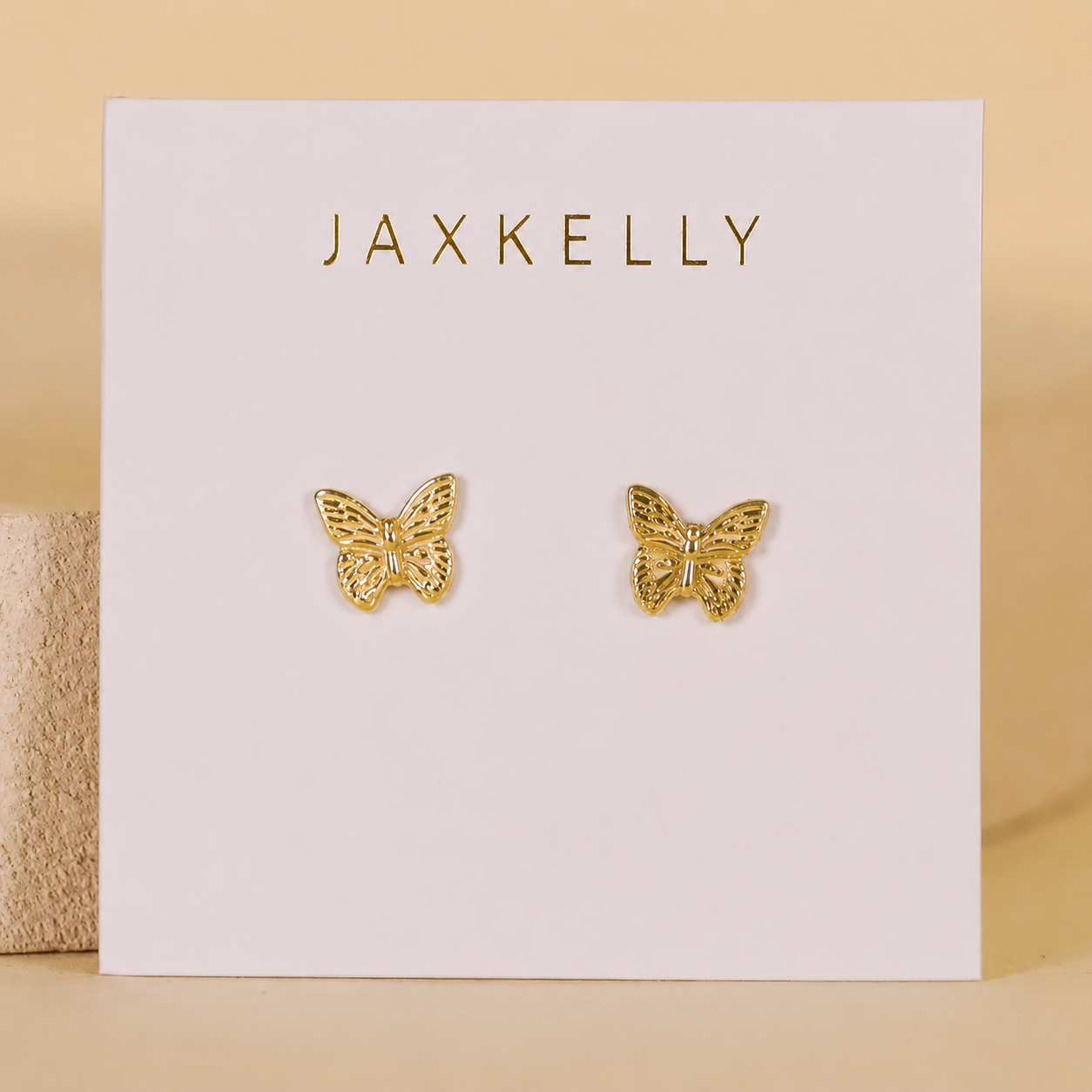 A pair of gold-plated Butterfly stud earrings by JaxKelly with sterling silver posts, shown on a white card with the JaxKelly logo. A soft beige background highlights this elegant jewelry.