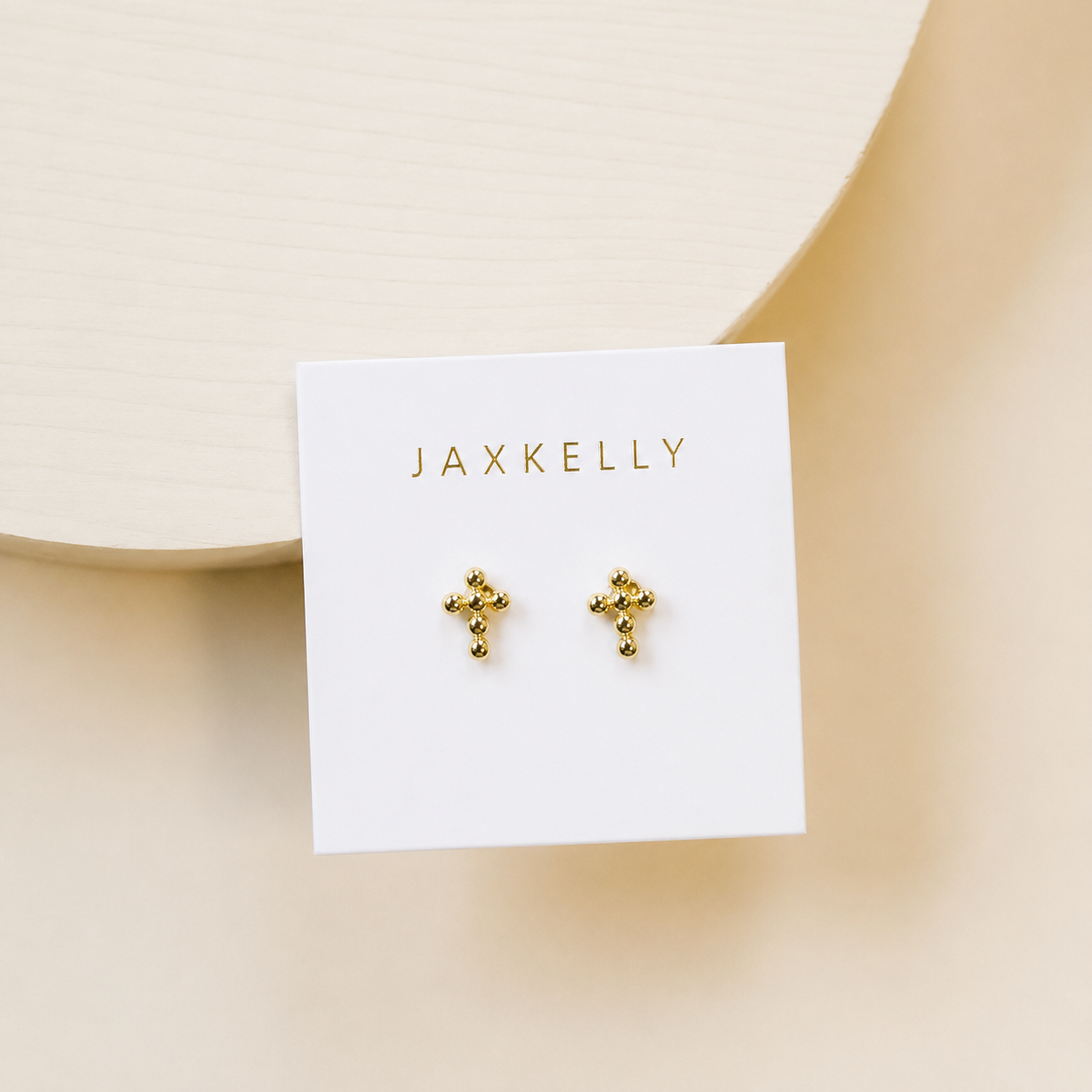 A pair of JaxKelly Cross Stud earrings with 18k gold plating and sterling silver posts rests on a white card labeled “JaxKelly” against a light neutral background.