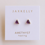 A pair of triangular amethyst stud earrings with 18k gold plating and gold prongs from JaxKelly, displayed on a white card labeled “Amethyst Mini Energy Gems - Healing.”.