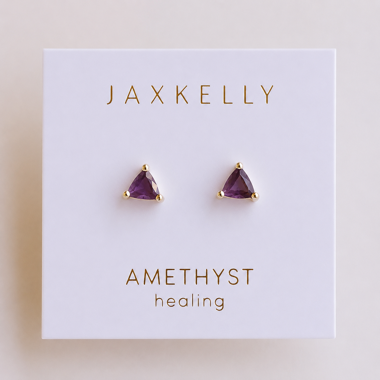 A pair of triangular amethyst stud earrings with 18k gold plating and gold prongs from JaxKelly, displayed on a white card labeled “Amethyst Mini Energy Gems - Healing.”.