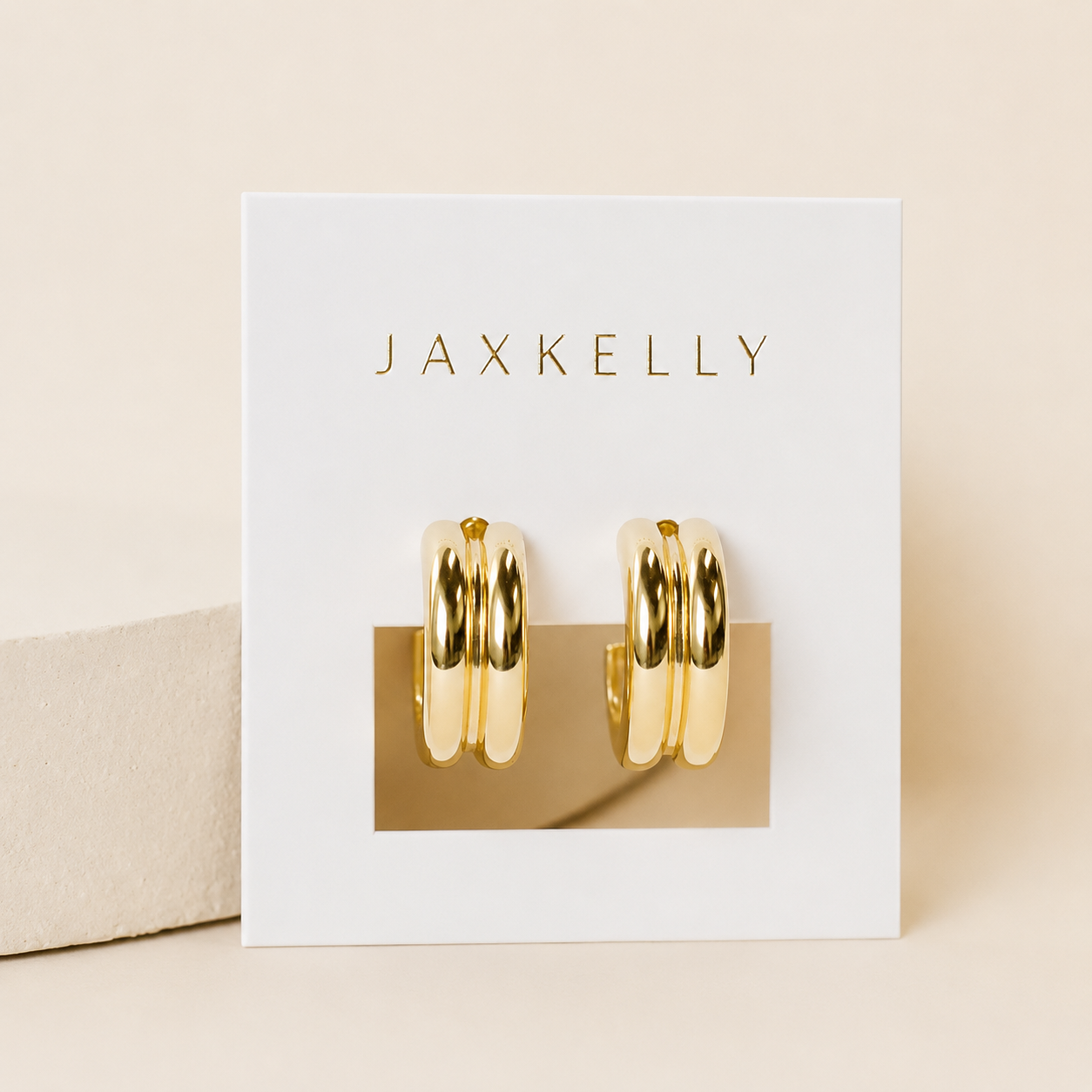 A pair of Bold Hoop earrings by JaxKelly, featuring shiny gold hoops with sterling silver posts, displayed on a white card with JaxKelly in gold lettering against a soft beige background.