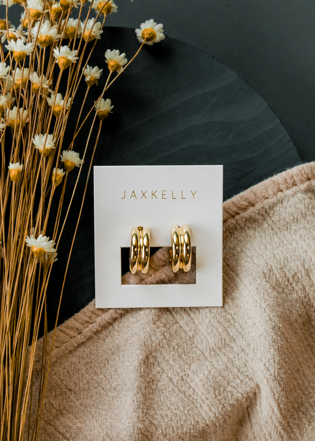 The JaxKelly Bold Hoop earrings—with sterling silver posts—rest on a white JaxKelly card beside dried yellow and white flowers and beige fabric atop a dark surface, making them the perfect long-lasting jewelry piece.