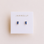 The Sapphire Baguette earrings by JaxKelly feature rectangular blue baguette studs set in gold-plated earrings with sterling silver posts, displayed on a white card with JaxKelly printed in gold letters.