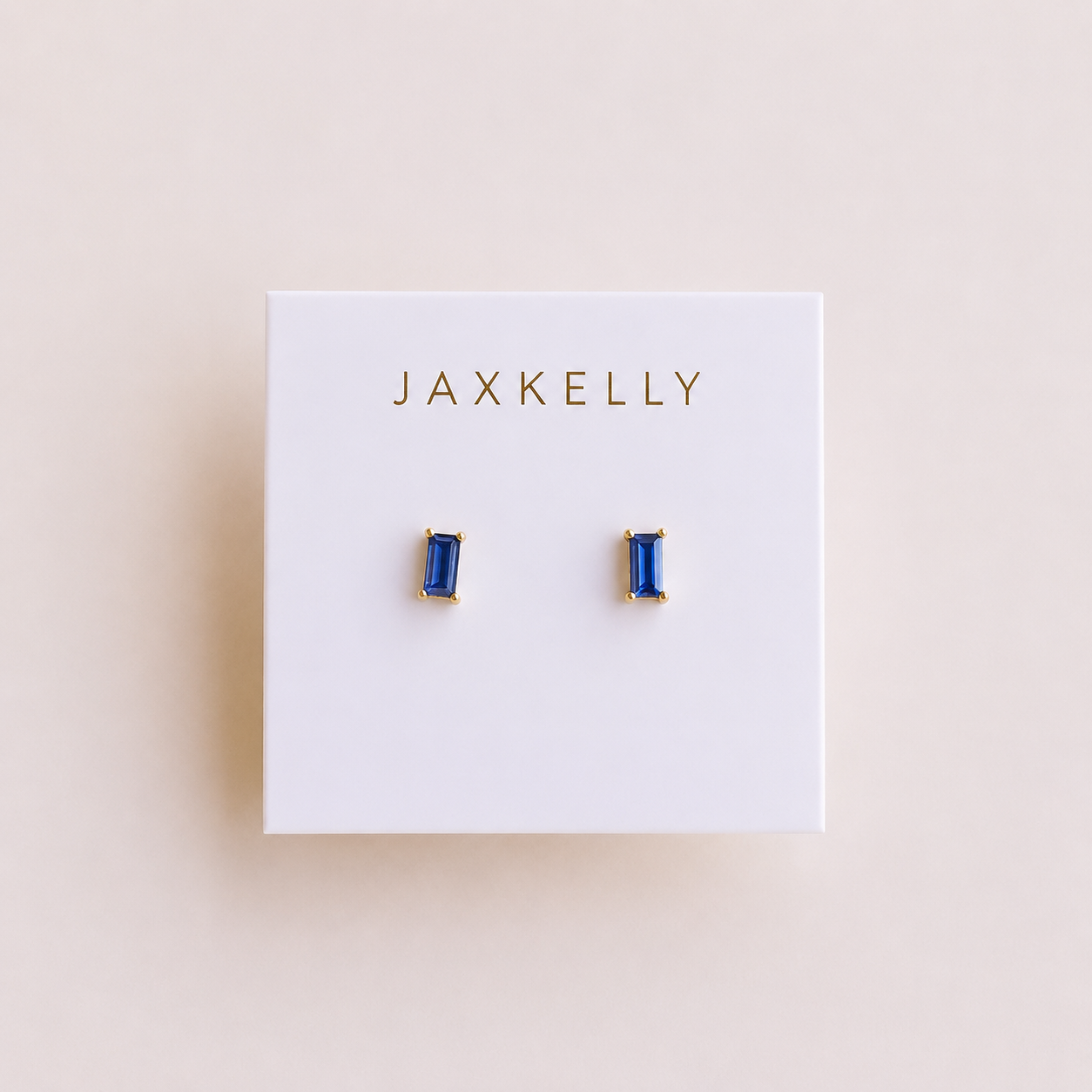 The Sapphire Baguette earrings by JaxKelly feature rectangular blue baguette studs set in gold-plated earrings with sterling silver posts, displayed on a white card with JaxKelly printed in gold letters.