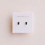A pair of JaxKelly Emerald Baguette stud earrings with rectangular green gemstones in gold settings and sterling silver posts, displayed on a white JaxKelly card against a light beige background.