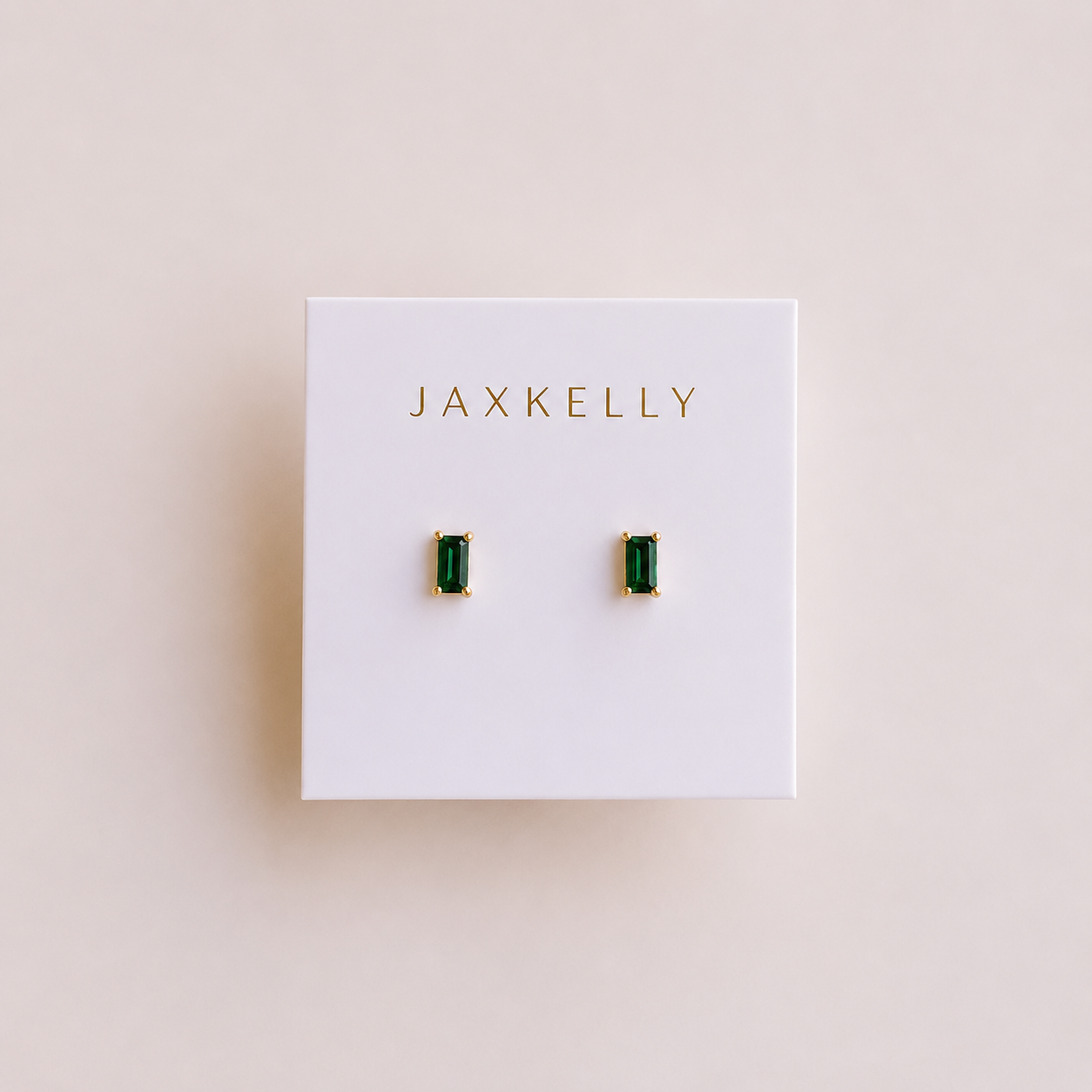 A pair of JaxKelly Emerald Baguette stud earrings with rectangular green gemstones in gold settings and sterling silver posts, displayed on a white JaxKelly card against a light beige background.