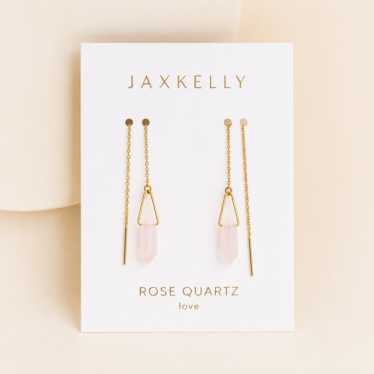 A pair of JaxKelly Gemstone Threader - Rose Quartz earrings with gold chains and rose quartz pendants, displayed on a white love card against a soft beige background.