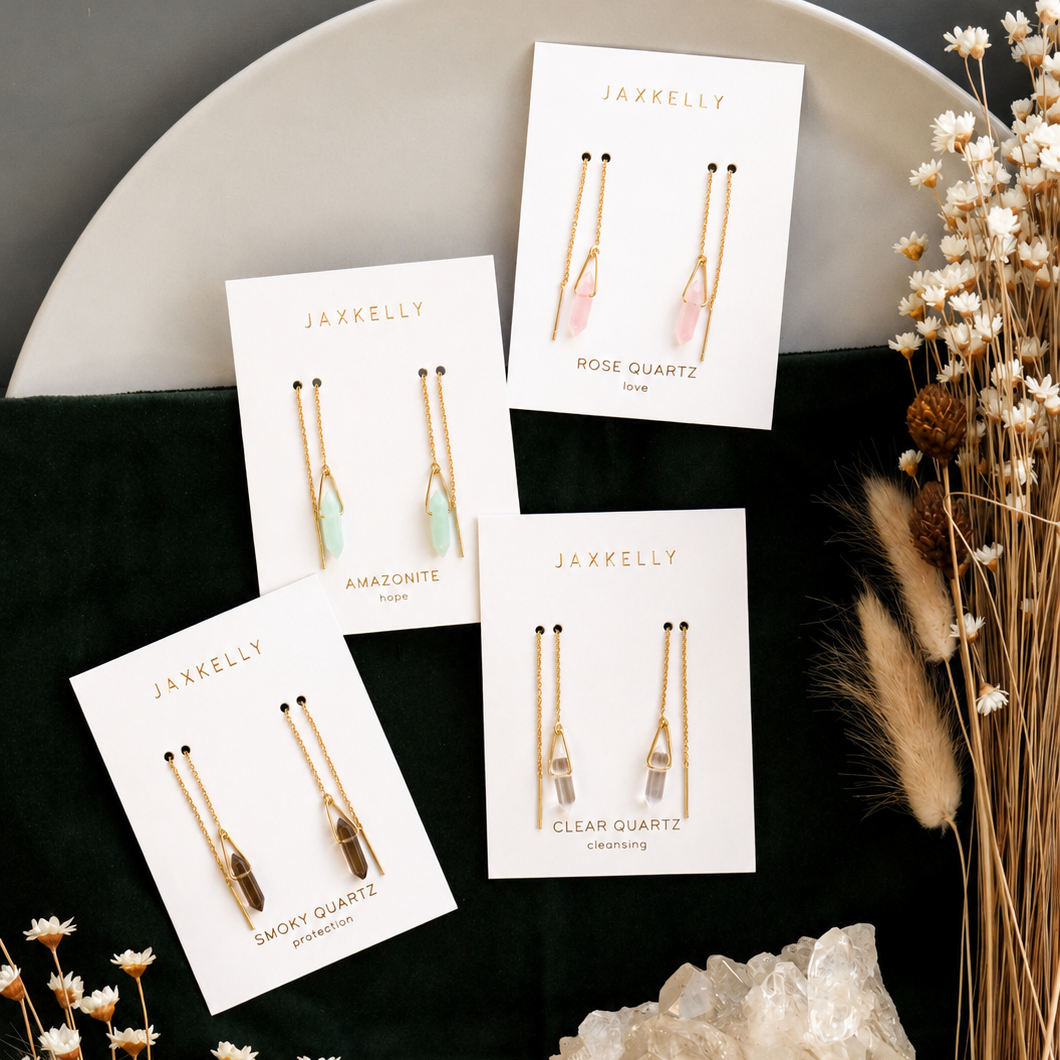 Four pairs of JaxKelly gemstone threader earrings, including Rose Quartz, Amazonite, Smoky Quartz, and Clear Quartz styles, are arranged on branded cards atop a round plate with dried flowers and a large clear crystal.