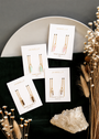 Four pairs of JaxKelly gemstone threader earrings, including Rose Quartz, Amazonite, Smoky Quartz, and Clear Quartz styles, are arranged on branded cards atop a round plate with dried flowers and a large clear crystal.