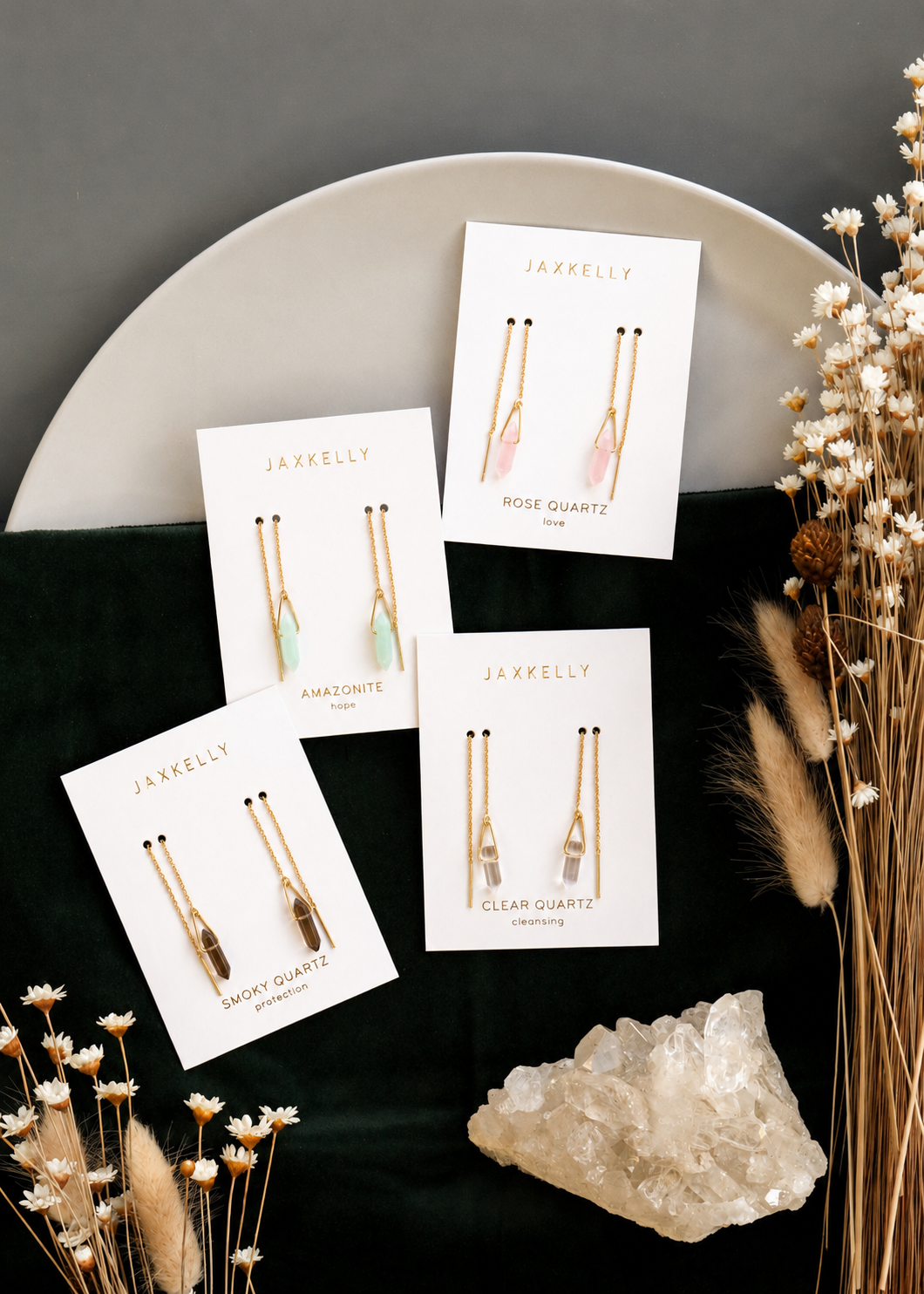 Four pairs of JaxKelly gemstone threader earrings, including Rose Quartz, Amazonite, Smoky Quartz, and Clear Quartz styles, are arranged on branded cards atop a round plate with dried flowers and a large clear crystal.
