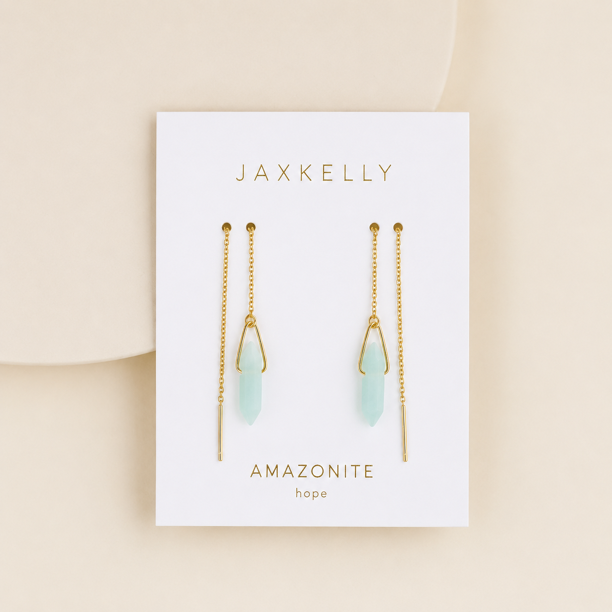 A pair of JaxKelly Gemstone Threader earrings featuring light green Amazonite crystal drops, presented on a white card labeled "JaxKelly" and "Amazonite hope," set against a soft beige background.