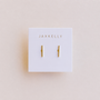A pair of Minimalist Bar earrings by JaxKelly, displayed on a white card with the brand name in gold at the top. Hypoallergenic design shown against a soft light beige background.