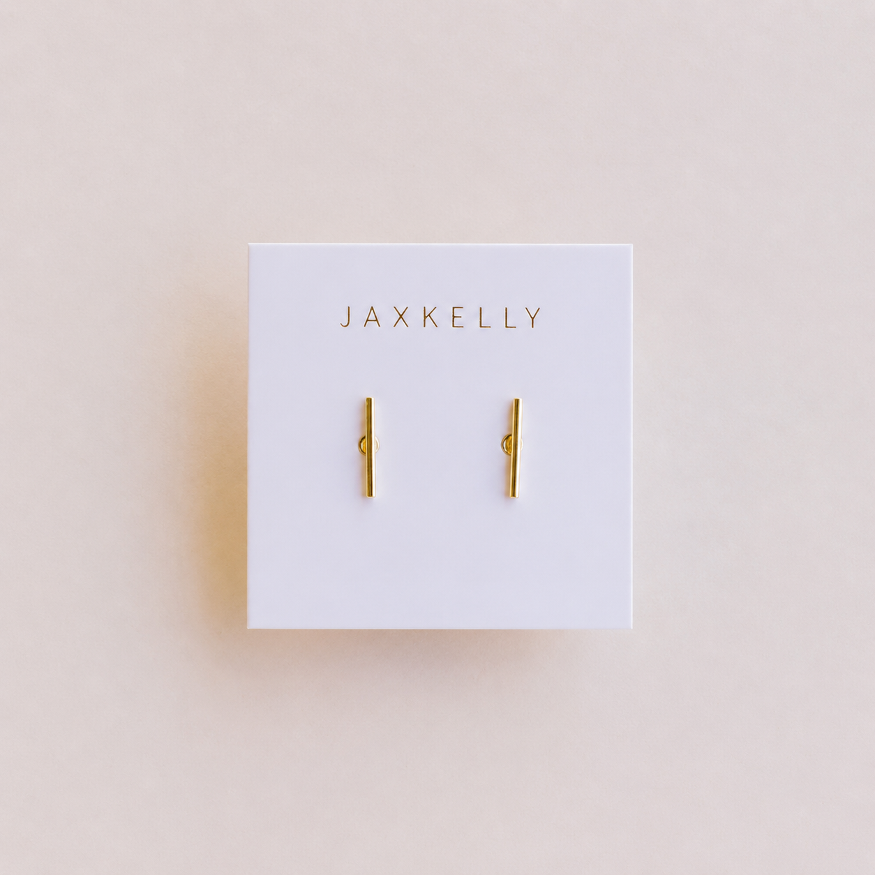 A pair of Minimalist Bar earrings by JaxKelly, displayed on a white card with the brand name in gold at the top. Hypoallergenic design shown against a soft light beige background.