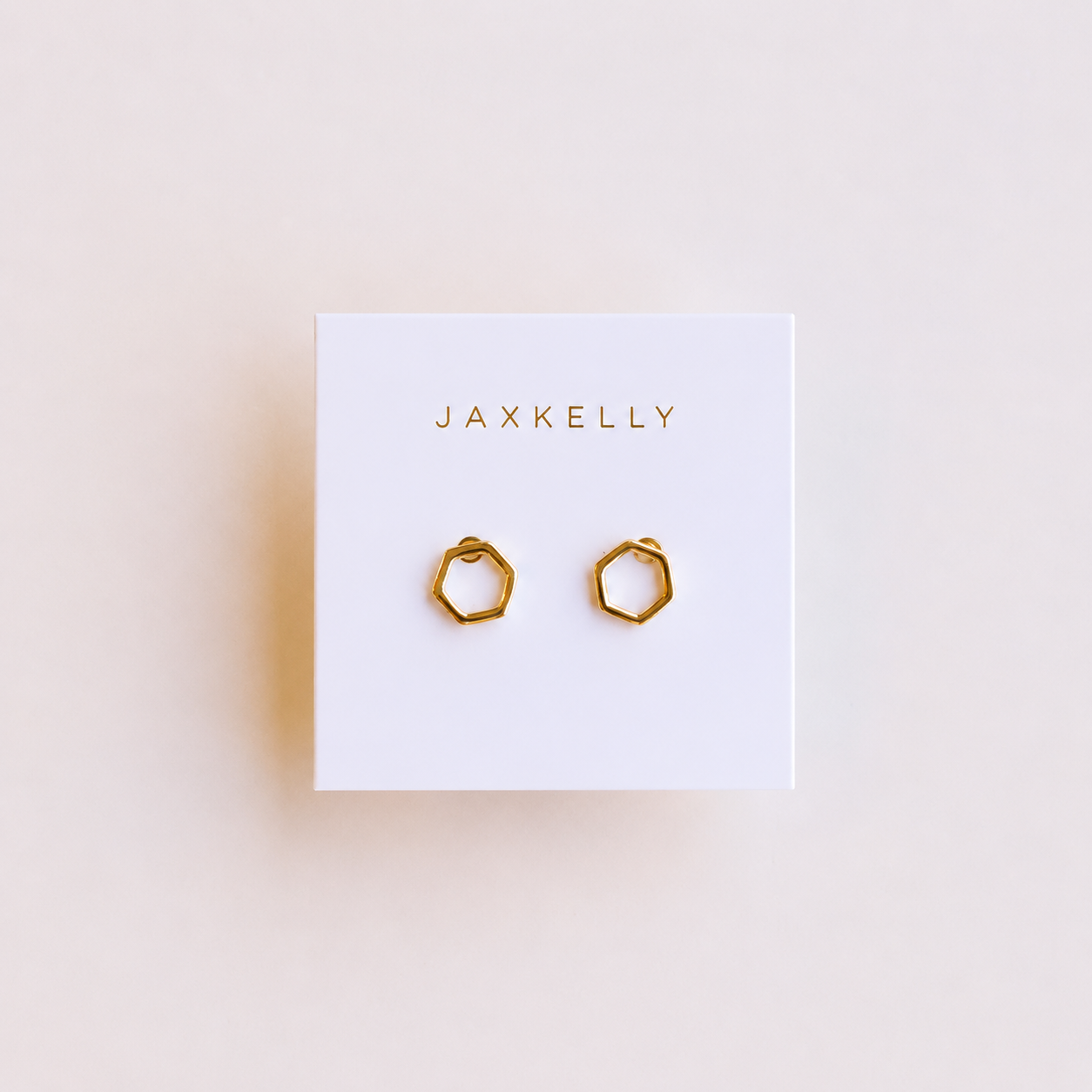 A pair of gold-plated Minimalist Hexagon earrings by JaxKelly displayed on a white card with JaxKelly in gold at the top; these hypoallergenic studs blend style and comfort.