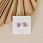 JaxKelly Amethyst Gemstone Prong - Healing stud earrings are shown with gold prongs on a branded card labeled "Amethyst healing," set against a beige background with a folded paper fan. These hypoallergenic earrings add a touch of natural beauty.