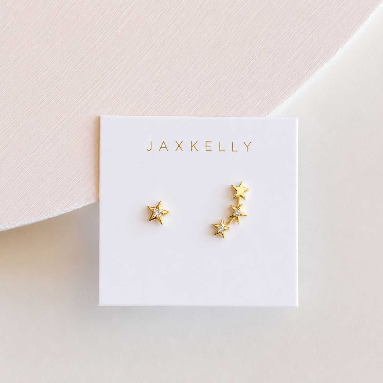 JaxKelly’s Star & Constellation earrings feature one large star on the left and a cascade of five smaller stars on the right, 18k gold plated and displayed on a white card.